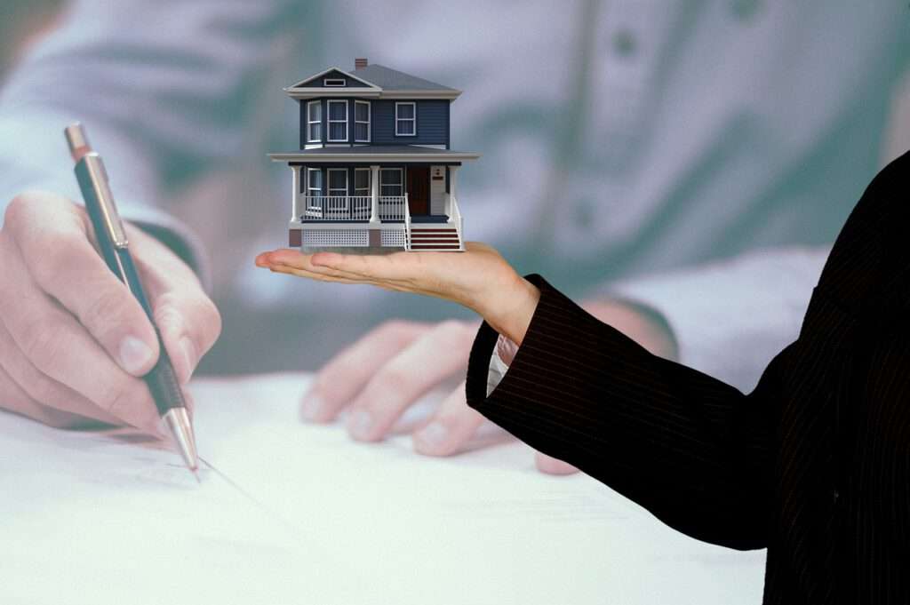 Im Vordergrund des Bildes ist eine Frauenhand, die ein kleines Modellhaus auf der Hand hält. Im Hintergrund, etwas weichgezeichnet sieht man eine Hand mit einem Kugelschreiber, die etwas auf ein Stück Papier schreibt.