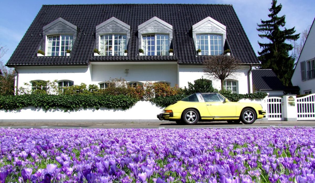 Eine tolle Villa in weiß mit einem gelben Porsche vor der Türe. Im Vordergrund befinden sich lila Blumen.