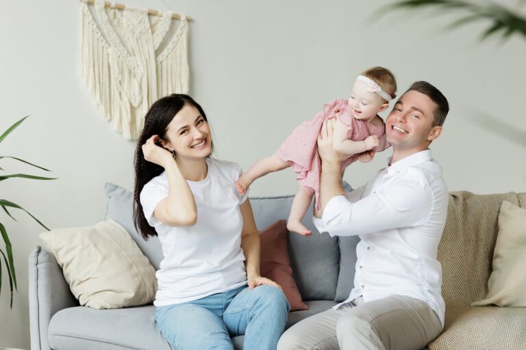 Auf dem Bild sieht man ein Mann, eine Frau mit Ihrem 2jährigen Kind auf einer Couch sitzen. Im Hintergrund ist eine graue Wand mit einem sandfarbenen Bild zu sehen.