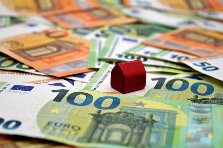 Auf dem Bild sieht man mehrere grüne Hundert Euro Scheine und ein paar braune 50 Euro scheine im Hintergrund. Mitten im Bild ist ein kleines rotes Holzhaus, welches auf einem Hudert Euro Schein steht zu sehen.