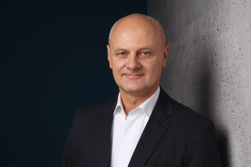 Professionelles Porträt eines Mannes in formeller Kleidung vor einer grauen Wand, lächelnd und selbstbewusst.